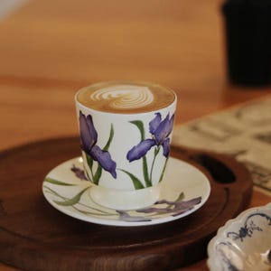 May include: A white ceramic coffee cup and saucer set featuring a purple and green iris floral design. The cup is filled with a latte, decorated with a heart-shaped foam pattern. The set rests on a wooden tray.