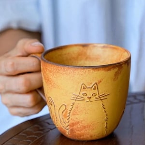 May include: A yellow ceramic mug with a cat design. The mug has a handle and a textured, rustic appearance. The cat is etched into the side of the mug. The mug is being held by a person.