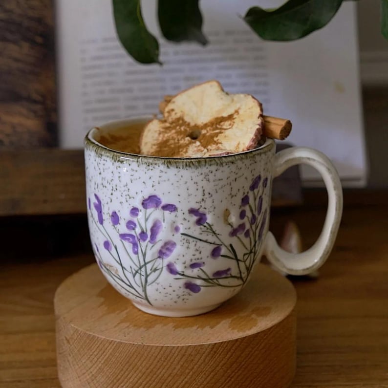 May include: A white ceramic mug with a floral design in purple, filled with a beverage and topped with a dried apple slice and cinnamon stick. The mug sits on a small wooden stand.