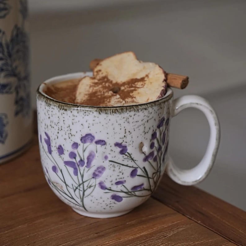 May include: A ceramic mug filled with a warm beverage, topped with apple slices, cinnamon, and a dusting of spice. The mug features a white speckled finish and a purple floral design. The mug is sitting on a wooden surface.