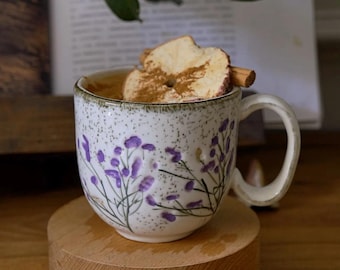 May include: A white ceramic mug decorated with purple floral patterns. The mug holds a warm drink, garnished with a dried apple slice and a cinnamon stick. It rests on a small wooden stand. The mug's handle is curved.