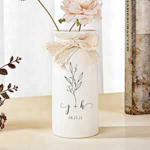 Puede incluir: Un jarrón de cerámica blanco con un diseño floral y un lazo de encaje. El jarrón tiene una ilustración botánica negra y las iniciales "j + h" con la fecha "08.23.25". Una flor rosa está en el jarrón.