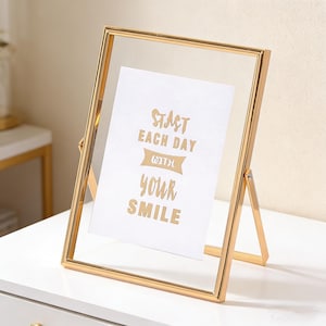 以下が含まれることがあります： 金色の金属製フォトフレーム、透明なガラスの前面。 フレームには、金色の文字で「Start Each Day With Your Smile」と書かれた白いカードが入っています。 フレームはスタンドで支えられています。