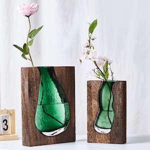 May include: Two decorative vases with a wooden frame and a green glass insert. One vase holds a pink rose, the other holds white flowers. The vases are on a white surface.