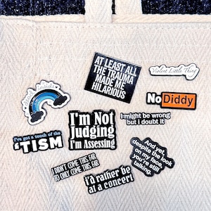 May include: A selection of enamel pins with humorous phrases and designs. The pins feature text such as "At Least All The Trauma Made Me Hilarious," "I'm Not Judging I'm Assessing," and "No Diddy." Other pins display a rainbow, clouds, and the words "Violent Little Thing."