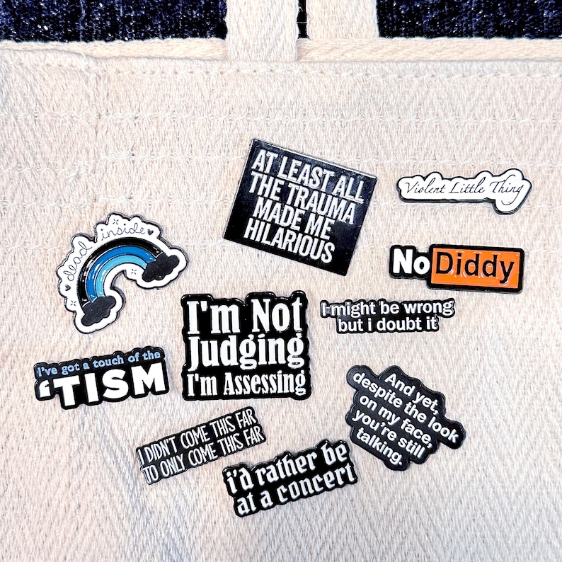 May include: A collection of colorful enamel pins with various phrases and designs. The pins include text like "At Least All The Trauma Made Me Hilarious," "I'm Not Judging I'm Assessing," and "No Diddy." Other pins feature a rainbow, clouds, and the words "Violent Little Thing."