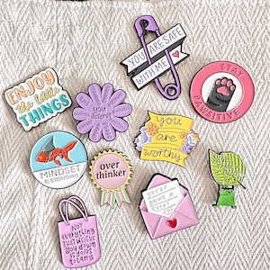 Op de afbeelding: Een verzameling kleurrijke emaille pins met diverse ontwerpen en tekst. Pins bevatten zinnen als "Enjoy the little things", "You are safe with me" en "Stay Pawsitive". Andere ontwerpen tonen een goudvis, een kikker en een envelop.