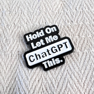 Könnte beinhalten: Ein schwarz-weißer Emaille-Pin mit dem Text "Hold On Let Me ChatGPT This." Der Pin hat eine rechteckige Form mit abgerundeten Ecken und ist vor einem strukturierten, cremefarbenen Hintergrund platziert.