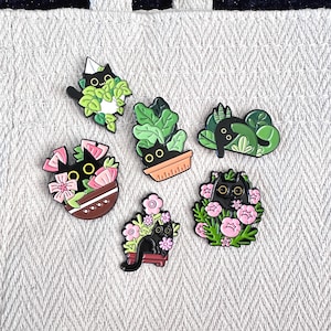 Puede incluir: Seis pines de esmalte con gatos negros en varios arreglos de plantas. Los pines muestran gatos rodeados de hojas verdes y flores rosas, con detalles como macetas y formas geométricas.