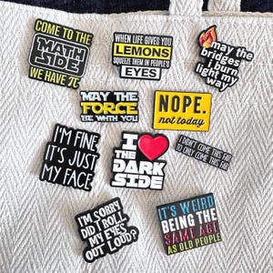 Könnte beinhalten: Eine Sammlung von schwarz-weißen Emaille-Anstecknadeln mit humorvollen Sprüchen. Die Anstecknadeln enthalten Zitate wie "Come to the Math Side", "When life gives you lemons" und "I'm fine, it's just my face". Die Anstecknadeln befinden sich auf einer strukturierten, cremefarbenen Oberfläche.