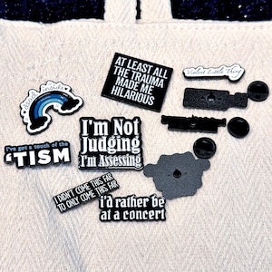 May include: A collection of enamel pins with various phrases and designs. The pins include text like "I'm Not Judging I'm Assessing", "At Least All The Trauma Made Me Hilarious", and "I'd Rather Be At A Concert". Other pins feature a rainbow and the words "dead inside".