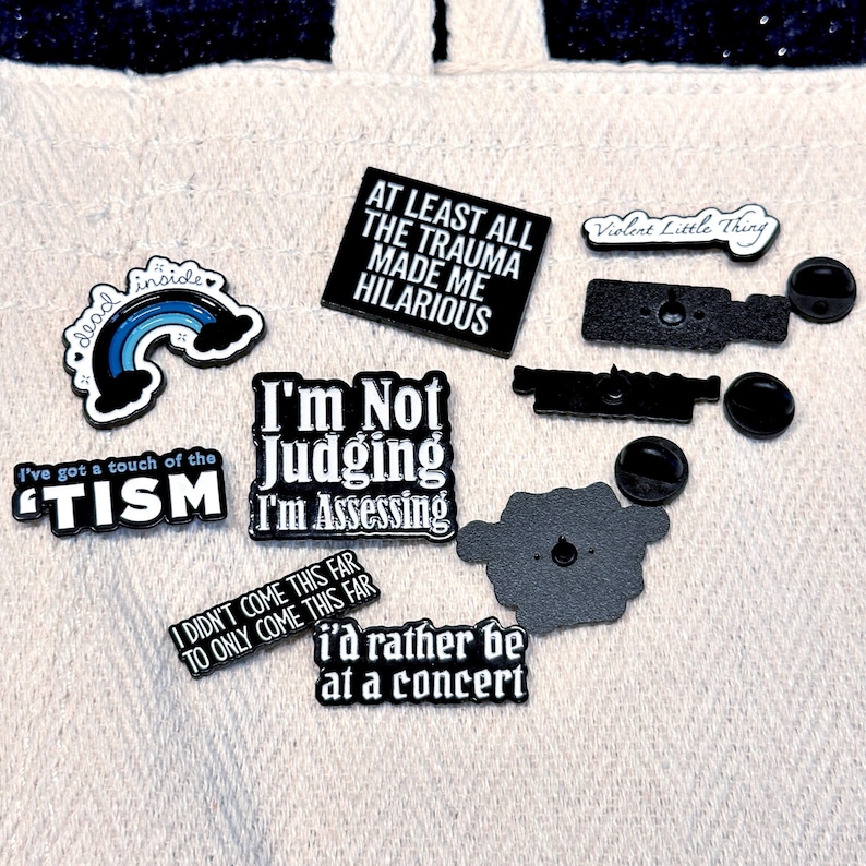 May include: A collection of enamel pins with various phrases and designs. The pins include text like "I'm Not Judging I'm Assessing", "At Least All The Trauma Made Me Hilarious", and "I'd Rather Be At A Concert". Other pins feature a rainbow and the words "dead inside".
