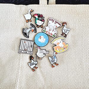 May include: A collection of whimsical goose-themed pins and patches. Designs include a goose in a cage, a goose with a lightsaber, and phrases like "Silly Goose Club" and "Have a Goose Day." The pins are set against a textured, off-white background.