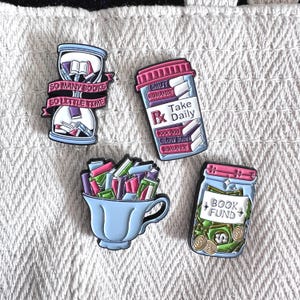 Peut inclure: Quatre breloques colorées sur le thème des livres. L'une est un sablier avec le texte "SO MANY BOOKS SO LITTLE TIME". Une autre est un flacon de pilules avec "Take Daily" et "ROMANCE". Une tasse est remplie de livres, et un bocal dit "BOOK FUND".