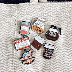 May include: A collection of coffee-themed enamel pins. Designs include a coffee pot with "Make Coffee Not War", a coffee cup, an IV bag with "100% Pure Coffee", and a pin that says "Hello I'm not caffeinated enough for this".