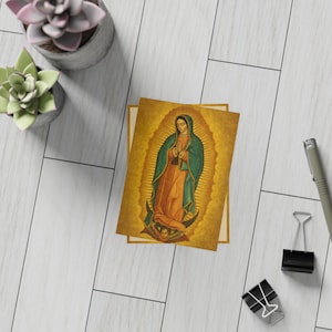 May include: A religious art print featuring the Virgin of Guadalupe. The image depicts a woman in a teal cloak and orange dress, surrounded by a golden halo. The print is on a cream-colored background.