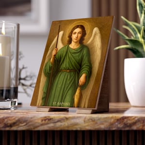 Puede incluir: Placa de madera con una imagen religiosa de San Rafael, representado con alas, un halo, sosteniendo un bastón y un pez. El santo viste una túnica verde. El texto "ST. RAPHAEL" es visible en la parte inferior.