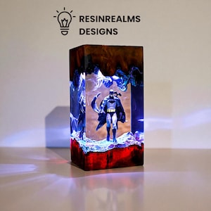 May include: A rectangular resin art piece featuring a Batman figurine. The piece has a layered design with brown, blue, and red hues. The text "RESINREALMS DESIGNS" is visible at the top. The piece is illuminated with internal lighting.