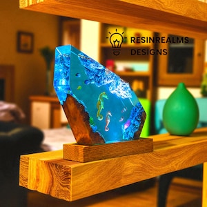 May include: A unique resin art piece featuring a vibrant underwater scene with seahorses and fish. The artwork is encased in a clear, blue resin block, mounted on a wooden base. The text "RESINREALMS DESIGNS" is visible.