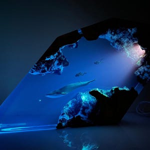 Puede incluir: Una obra de arte de resina azul que representa una escena submarina con una ballena, buzos y formaciones rocosas. La obra está iluminada con luz azul, creando un efecto dramático. La ballena nada en el centro de la pieza.