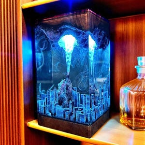 May include: A blue-lit resin sculpture of Godzilla in a city setting. The rectangular piece has a dark, textured base and top. The sculpture's interior is illuminated, with a bright light source at the top, creating a striking visual effect.
