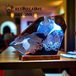 May include: A unique resin art piece featuring a dragon silhouette and mountain landscape. The artwork is encased in a clear, geometric form with a wooden base. The piece has a blue and brown color scheme, with the text "RESINREALMS DESIGNS".