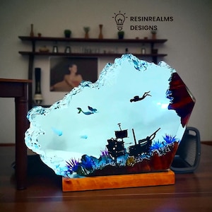 Könnte beinhalten: Dekorative Harzskulptur mit einer Unterwasserszene mit einem gesunkenen Schiff, einem Taucher und Meereslebewesen. Die Skulptur hat einen hellblauen, strukturierten Rand und steht auf einem Holzsockel. Der Text "RESINREALMS DESIGNS" ist sichtbar.