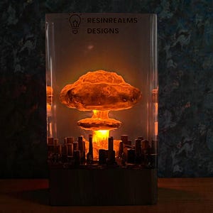 May include: A rectangular resin lamp featuring a glowing orange mushroom cloud design above a cityscape. The lamp has the text "RESINREALMS DESIGNS" and a lightbulb icon. The lamp is lit from within, creating a warm, ambient glow.