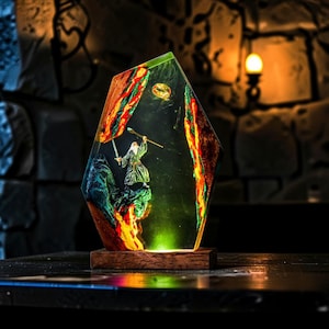 Può includere: Una lampada decorativa in resina e legno con una miniatura di un mago che impugna un bastone e una spada, posizionata all'interno di un blocco traslucido dai bordi irregolari. L'oggetto include un anello dorato sospeso sopra la figura e una base luminosa.