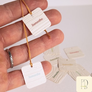 Puede incluir: Una colección de pequeñas etiquetas cuadradas de color blanquecino con textura de vetas de madera. Cada etiqueta tiene el nombre "Samantha" impreso en un color suave. Las etiquetas están unidas a finos hilos dorados.