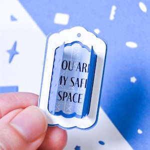 May include: A blue and white paper craft with the words "YOU ARE MY SAFE SPACE" printed on it. The craft is held by a person's fingers against a blue background with white shapes.