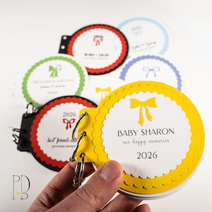 May include: A yellow baby memory book with a scalloped edge and a yellow bow design. The cover reads "BABY SHARON our happy memories 2026." Other pages feature bow designs and names. The book is held together with metal rings.
