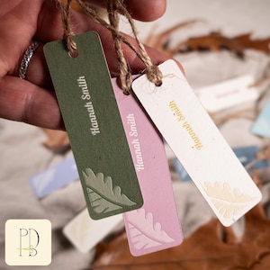 Può includere: Primo piano di tre etichette regalo rettangolari verdi, rosa e bianche, ciascuna con il nome "Hannah Smith" stampato verticalmente. Le etichette presentano un disegno a foglia e sono attaccate a spago. Lo sfondo include altre etichette e foglie autunnali.