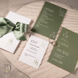 May include: Wedding invitation suite featuring olive green and white cards. Includes an invitation, RSVP card, and details card. A satin green ribbon and two gold rings are also present. The suite is arranged on a beige textured surface.