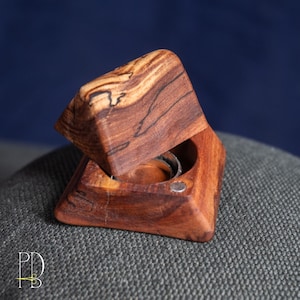 May include: A handcrafted wooden ring box with a hinged lid, showcasing a rich brown color with dark grain patterns. The box is open, revealing a silver ring inside. The box is set against a dark blue backdrop.