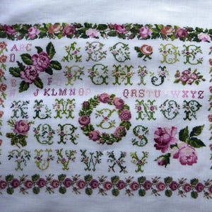 Può includere: Campione a punto croce con alfabeto e motivi floreali. Le lettere sono ricamate in verde e rosa, circondate da rose e foglie. Il ricamo ha un bordo floreale su sfondo bianco.