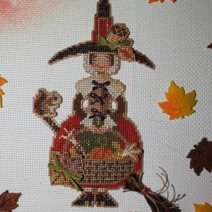 May include: A cross-stitch embroidery of a figure wearing a tall pointed hat, a white collar, and a red skirt, holding a basket filled with autumn produce. The figure is accompanied by a small squirrel and a broom with brown tassels.