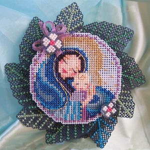 Può includere: Un ricamo a punto croce circolare che raffigura una figura stilizzata che tiene in braccio un bambino, incorniciato da foglie perforate grigio scuro con cuciture verdi. L'immagine centrale presenta fili blu, oro e rosa, accentuati da piccoli fiori in nastro.