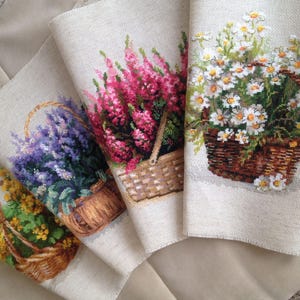 Può includere: Quattro pezzi di tessuto con disegni floreali a punto croce. Ogni pezzo presenta un cestino pieno di fiori in vari colori: giallo, viola, rosa e bianco. I cestini sono marroni, su uno sfondo beige neutro.