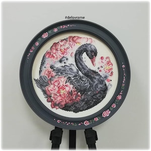 May include: A framed cross-stitch artwork featuring a black swan surrounded by pink peonies. The artwork is in a round, gray frame with floral accents. The text "#delovrame" is in the upper left corner.