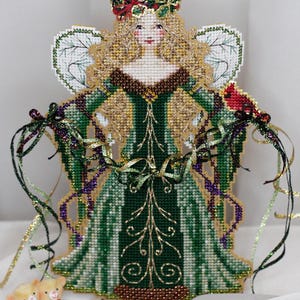 May include: A cross-stitch angel figurine with blonde hair, wearing a dark green gown with gold embroidery. The angel has white wings, a holly crown, and holds a red cardinal. Two small teddy bears with a heart are positioned at the base.