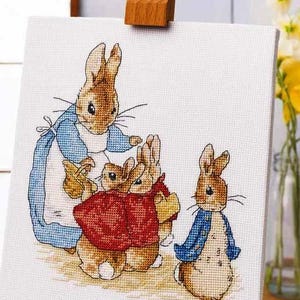 May include: Cross-stitch embroidery of a rabbit family, featuring a mother rabbit in a blue dress and three smaller rabbits. The rabbits are in shades of brown, red, and blue. The artwork is displayed on a small wooden easel.