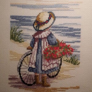 May include: A framed cross-stitch embroidery depicting a person wearing a straw hat, a blue dress, and a white shawl, standing on a sandy path with a bicycle. A basket filled with red flowers is attached to the bicycle, with a blue sea in the background.