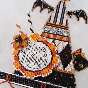 May include: A decorative Halloween-themed cross-stitch piece featuring a tall, striped hat topped with a jack-o'-lantern. It includes a circular sign reading Happy Halloween, a black cat silhouette, bat wings, and orange and black beaded accents.