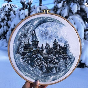 Può includere: Ricamo a punto croce in un telaio di legno raffigurante una scena di castello sotto una luna crescente. L'opera presenta un castello dettagliato con più torri, circondato da alberi innevati, nelle tonalità del blu e del bianco.