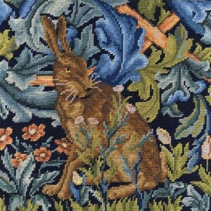 Può includere: Un arazzo a punto croce raffigurante una lepre marrone al centro di un fitto fogliame. Il disegno include foglie blu scuro in stile acanto, fiori arancioni vivaci e piccoli fiori bianchi su uno sfondo scuro, realizzato a punto croce.