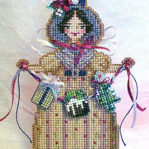 Könnte beinhalten: Ein Kreuzstich-Ornament in Form einer Person mit blauer Haube und beigem Kleid mit vertikalen Streifen. Die Figur hält Bänder, an denen kleine Kreuzstich-Anhänger befestigt sind, darunter ein Geschenk, ein Pudding und ein grüner Baum.
