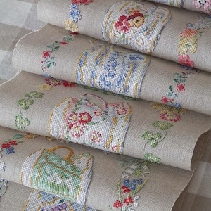 Puede incluir: Primer plano de varias piezas de tela de lino enrolladas. Cada pieza presenta diseños florales bordados a punto de cruz en rojo, azul y amarillo. La tela tiene un tono beige neutro, y los diseños están dispuestos en un patrón repetitivo.