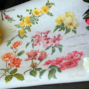 Puede incluir: Un bordado de punto de cruz detallado con diferentes variedades de rosas en tonos amarillos, rosas y rojos. El bordado incluye nombres botánicos como "Canary Bird" y "Sea Pearl" sobre tela color crema.