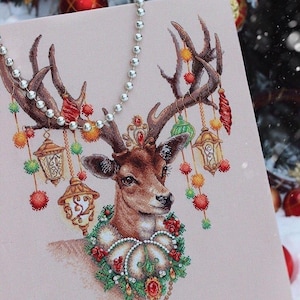 Può includere: Un disegno a punto croce con una renna con corna ornate di decorazioni natalizie e lanterne. La renna indossa una corona di gioielli e una ghirlanda festiva. Una ghirlanda di perline d'argento è appesa sopra l'immagine.